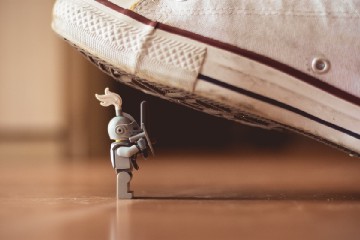A Lego figure sits on the floor below a shoe, which threatens to crush it